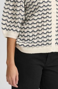 Cardigan en tricot de couleur crème avec un motif en vagues bleu marine et marron, doté d'une fermeture à boutons et de poignets et d'un ourlet côtelés.