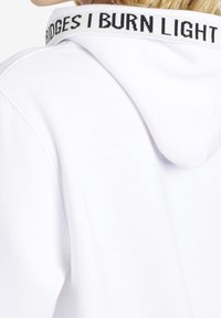 Ingrandimento della parte posteriore di un hoodie bianco con cappuccio alzato, caratterizzato da un colletto con una rifinitura nera su cui è scritto "BRIDGES I BURN LIGHT".