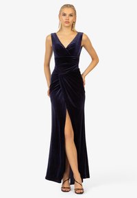 Nachtblaues Samt-Abendkleid mit V-Ausschnitt, figurbetonter Taille und seitlichem Schlitz. Die drapierte Details an der Taille betonen die elegante Silhouette.