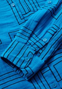 Camisa azul de botones con patrones lineales negros, textura suave y puño abotonado con un detalle de botón azul claro.
