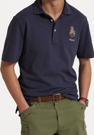 Mężczyzna ubrany w granatową koszulkę polo z haftowanym logo niedźwiedzia oraz oliwkowe spodnie typu cargo z brązowym paskiem i zegarkiem, z rękami w kieszeniach.