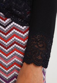 Černá krajková manžeta s jemnými květinovými vzory, doplněná vícebarevným látkovým vzorem se zubatými pruhy v červené, bílé a černé barvě.