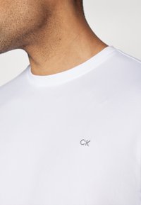 T-shirt branca de algodão com decote redondo e um pequeno logótipo da CK bordado em cinza no peito, com uma textura suave.