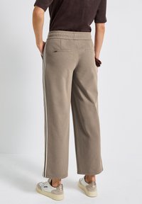 Beige, weit geschnittene Sweatpants aus weichem Material, mit seitlichen Streifen, elastischem Bund und zwei Gesäßtaschen. Kombiniert mit lässigen Sneakers.