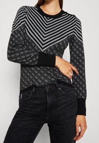 Schwarzer Pullover mit geometrischen Streifen und Rautenmustern, rundem Halsausschnitt und gerippten Bündchen, getragen mit dunklen Jeans.