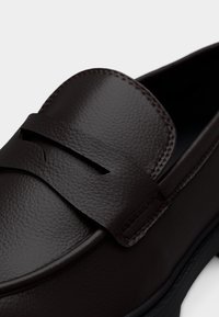 Σκούρο καφέ δερμάτινο μοκασίνι με υφασμένη επιφάνεια, διακοσμητική λωρίδα penny, στρογγυλεμένη μύτη και εμφανείς ραφές κατά μήκος των άκρων.