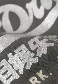 Tessuto grigio con testo in glitter metallico argento. Le lettere hanno dimensioni e forme diverse, creando un aspetto tridimensionale rispetto allo sfondo.