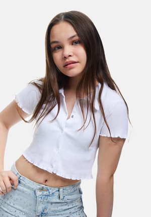 Witte geribbelde crop top met korte mouwen en een golvende zoom, met een kleine knoopsplit. Gecombineerd met lichtblauwe versierde jeans.