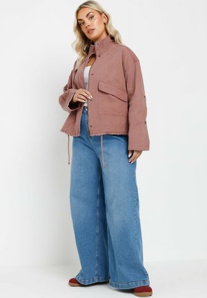 Giacca rosa tagliata con colletto alto, tasca sul petto e orlo sfilacciato, abbinata a pantaloni in denim blu a gamba larga e scarpe rosse.
