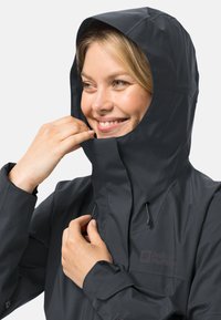 Zwarte waterdichte jas met een aansluitende capuchon, voorzien van een ritssluiting aan de voorkant, aantrekkoorden en een gestructureerde stof. Logo zichtbaar op de borst.