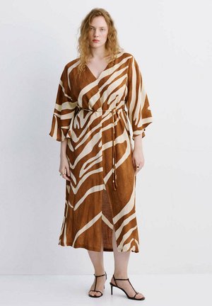 Mujer con cabello ondulado, vestida con un vestido cruzado de rayas de cebra marrón y beige, con mangas tres cuartos y tacones negros con tiras.