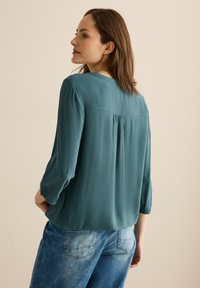 Teal bluse med lange ærmer, rygfold-detalje, glat tekstur. Parret med lyseblå denimjeans. Neutral baggrund.