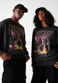 Sweat-shirt noir avec le graphique "CHICAGO BULLS", arborant le logo du taureau, des éléments de basket-ball et un fond enflammé. Associé à des casquettes noires assorties.