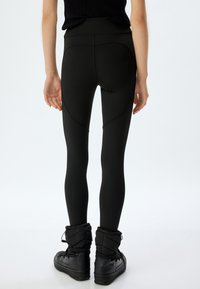 Zwarte hoge taillleggings met gebogen naaddetails, gemaakt van rekbaar materiaal. Gekleed met zwarte gewatteerde enkellaarsjes.