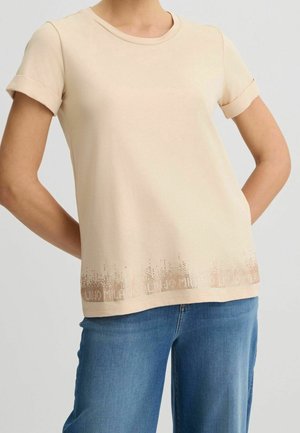 Person, der ein beige kurzärmliges T-Shirt mit einem gepunkteten Farbverlauf am Saum und eine blaue Jeans trägt, steht mit Händen auf dem Rücken.
