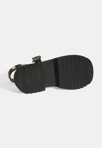 Black sandal with a textured rubber sole, featuring adjustable straps and round gold accents. Sole has deep grooves for traction.