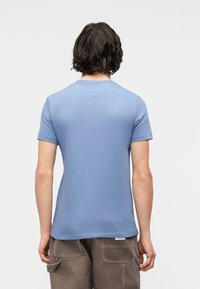 Άτομο με κοντά σκουρόχρωμα μαλλιά, φοράει ανοιχτό μπλε κοντομάνικο πουκάμισο και καφέ cargo παντελόνια, κοιτάζει προς τα πίσω σε μια μονόχρωμη σκηνή.