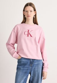 Felpa rosa con colletto rotondo, vestibilità ampia, con il logo "Calvin Klein" e "CK" in rosso scuro. Abbinata a jeans in denim blu.