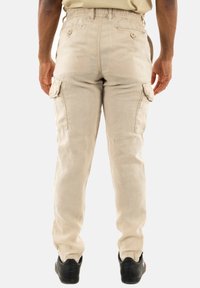 Beige cargobroek met een relaxte pasvorm, elastische tailleband en dubbele zijzakken. Gemaakt van lichtgewicht stof. Zwarte schoenen zichtbaar.