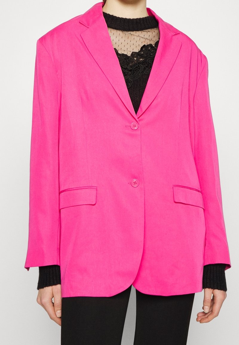 Femme portant un blazer rose vif à deux boutons sur un haut en dentelle et mesh noir avec un pantalon noir, vue du cou aux hanches.