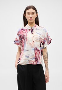 Mladá žena s dlouhými tmavými vlasy, oblečená v krátkým rukávem košili s abstraktními vzory v červené, růžové a modré barvě a černé kalhoty.