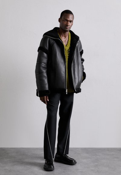 Black leather oversized jacket with a fur-lined collar, paired with a green knit sweater and black pants featuring white side piping. Black shoes.