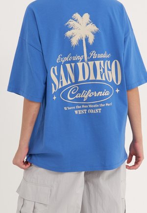 Niebieska oversize koszulka z beżową grafiką palmy i napisem "Exploring Paradise San Diego California" noszona z jasnymi szortami typu cargo.