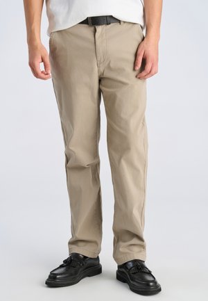 Man wearing beige chinos, black leather tassel loafers, black belt, and white shirt, standing against a plain light background.