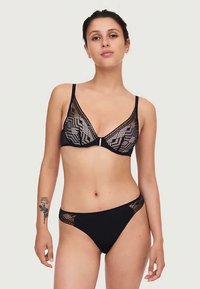 Γυναίκα φορώντας μαύρο σύνολο εσωρούχων από δαντέλα με γεωμετρικό μοτίβο στις θηλές και λεπτομέρειες από πλέγμα στα σλιπ, που στέκεται μπροστά σε απλό φόντο.