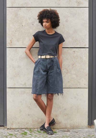 Sort T-shirt, høj talje mørke denim shorts med frayed kant, fastgjort med et beige bælte med nitter, og sorte slip-on sko med udskæringer.