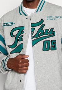 Giacca varsity grigia con strisce teal. Grande grafica "FUBU" in teal e nero sul davanti, con bottoni e polsini a coste.