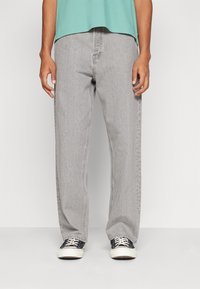 Osoba ve volných světle šedých džínách, černobílých teniskách a zkráceném zeleném tričku na jednobarevném bílém pozadí.