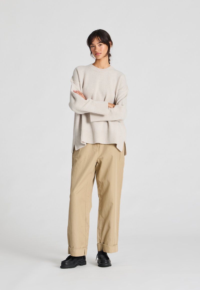 Licht beige oversized gebreide trui met ribbelmanchetten, gecombineerd met iets losse tan cargobroek en zwarte veterschoenen.