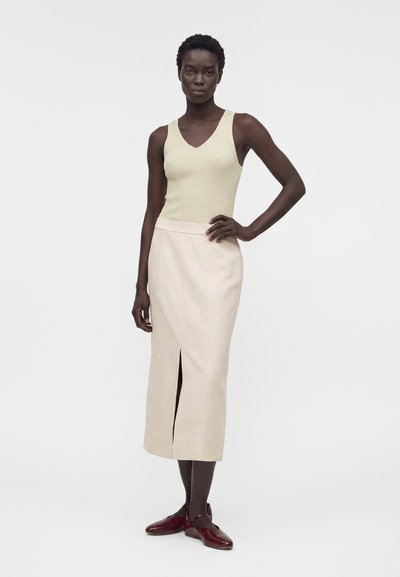 Γυναίκα που φοράει μπεζ μπλουζάκι χωρίς μανίκια με ανάγλυφη υφή, κρεμ midi φούστα με σκίσιμο μπροστά και σκούρα κόκκινα παπούτσια Mary Jane, στέκεται με το ένα χέρι στη μέση.