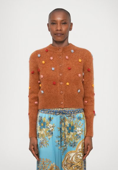 Cardigan en peluche marron avec des pompons multicolores en rouge, jaune, bleu et rose ; fermeture à boutons et col rond ; associé à une jupe bleue à motifs.