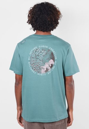 Personne aux cheveux bouclés portant un t-shirt bleu sarcelle avec un graphisme rond inspiré par la nature et un texte au dos.