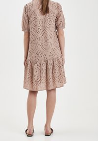 Světle růžové šaty z vyšívaného materiálu s několika vrstvami sukně, krátkými nabíranými rukávy a složitými perforovanými vzory po celém povrchu látky.