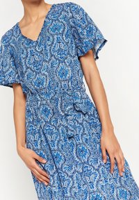Modré šaty se vzorem, které mají výstřih do V, krátké rukávy a zúžený pas, vyrobené z lehké tkaniny s paisley vzorem.