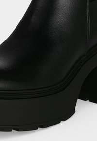 Gros plan d'une botte à plateforme en cuir noir avec talon épais et semelle texturée sur un fond clair.