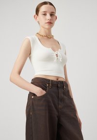 Hvit ribbet crop top med snøring i halsen, paret med høydejusterte mørkebrune jeans med kobberdetaljer og side lommer.