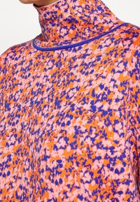 Stickad polotröja med en livlig orange bakgrund prydd med ett mönster av rosa och blå prickar samt lila kant vid kragen.