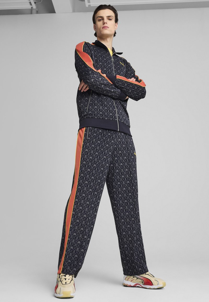 Tuta in fantasia navale con accenti arancioni e gialli; presenta una giacca con zip e pantaloni con vestibilità rilassata, abbinati a sneakers multicolori.