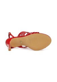 Sandalia roja de tacón alto con tiras, tumbada de lado mostrando la suela beige texturada con un pequeño logo dorado.