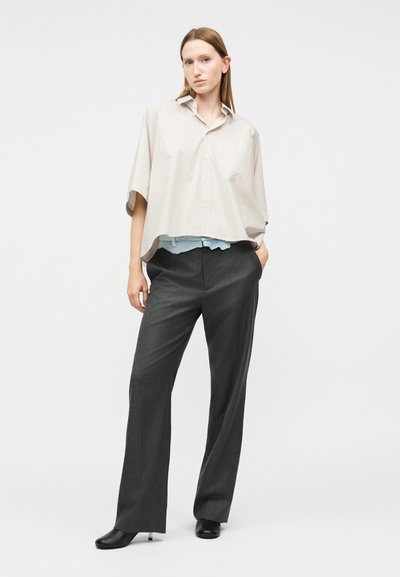 Kobieta o prostych włosach, ubrana w oversize'ową beżową koszulę z krótkim rękawem, ciemne szerokie spodnie oraz czarne buty na obcasie, stojąca z jedną ręką w kieszeni.