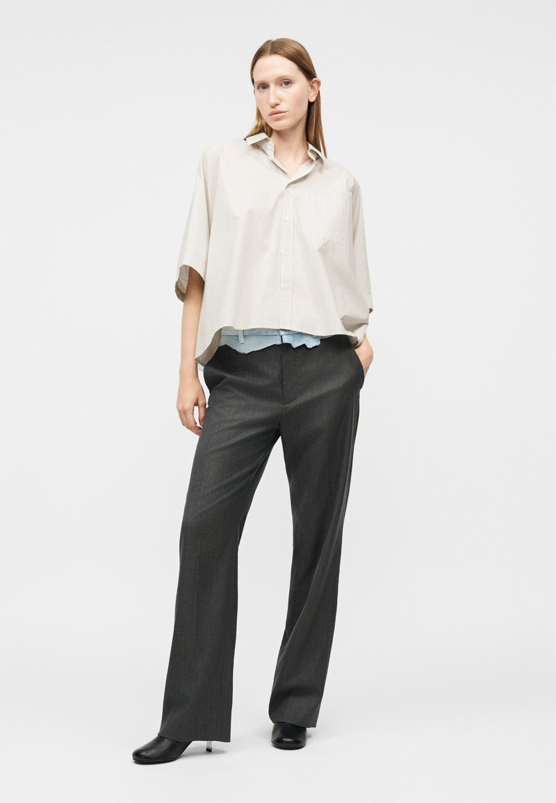 Vrouw met steil haar, gekleed in een oversized beige kortemouwenoverhemd, donkere losse broek met wijde pijpen en zwarte schoenen met hakken, staand met een hand in haar pocket.