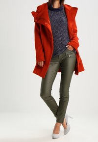 Cappotto rosso in lana con un ampio colletto, maglione blu navy testurizzato, pantaloni aderenti verde oliva e scarpe con tacco grigie.