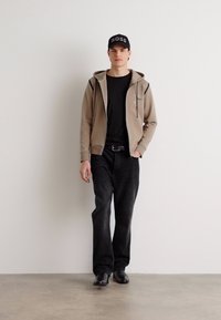 Haut à zip beige sur un t-shirt noir, associé à un jean noir et des chaussures noires. Le modèle porte une casquette noire avec le mot "BOSS" en blanc.