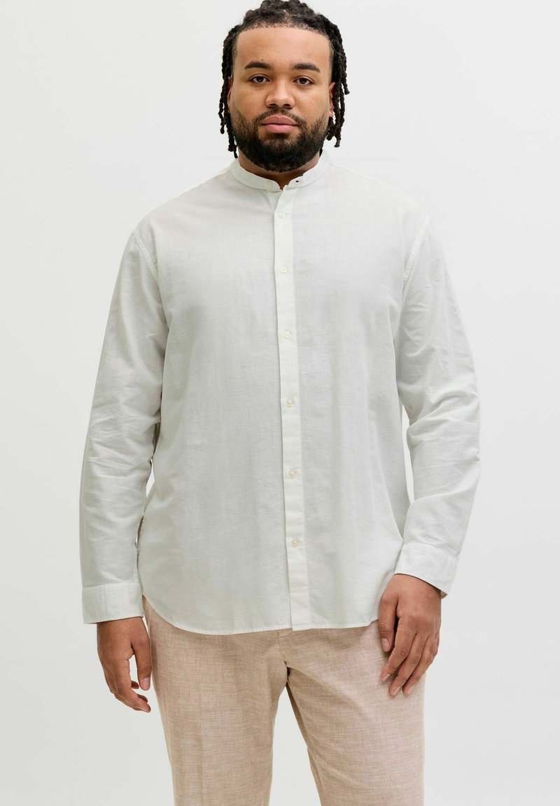 Homme aux dreadlocks courtes portant une chemise blanche à col mao boutonnée et un pantalon beige, debout devant un fond clair uni.