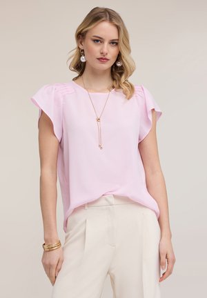 Blondynka w jasnoróżowej bluzce z plisowanymi rękawkami typu cap, białych spodniach z wysokim stanem, złotym naszyjnikiem, perłowymi kolczykami i złotymi bransoletkami na neutralnym tle.