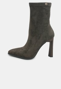 Stivale alla caviglia in suede marrone con punta appuntita, tacco a stiletto alto, cuciture rinforzate e un accento con logo vicino alla parte superiore. Texture liscia.